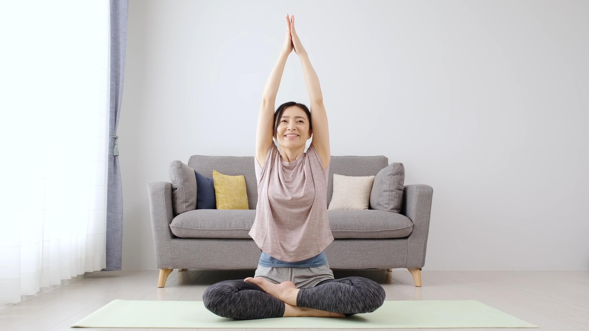 座禅を組みながら、合わせた両手を頭の上に伸ばす女性